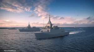Three modern naval ships sail on calm water under a partly cloudy sky at sunset. The ships are grey and equipped with radar and communication equipment. Distant land is visible on the horizon.