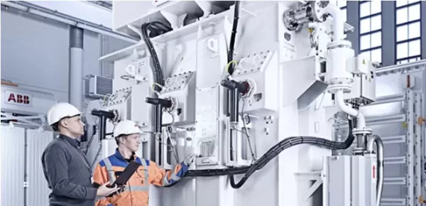 Two engineers wearing safety helmets and work uniforms inspect and discuss a large industrial electrical machine inside a bright, modern facility. One holds a tablet while both look at the equipment.
