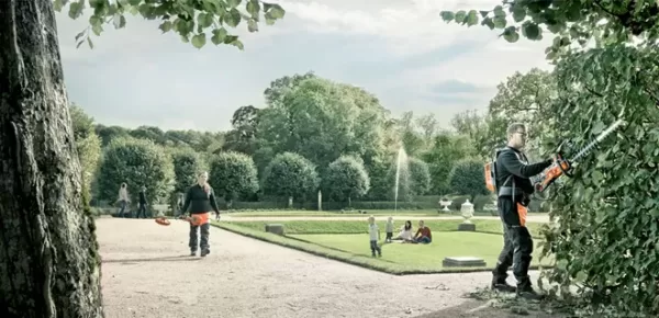 Several people are in a well-maintained park; one person trims a hedge with an electric trimmer, another walks with gardening tools, while others sit or walk on the grass and paths among green trees and shrubs.