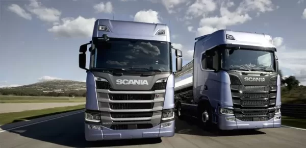 Two large Scania trucks, silver in color, are parked side by side on a road with a clear sky and hills in the background.