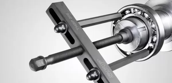 A close-up of a mechanical bearing puller tool attached to a metal bearing assembly, showing the threaded rods, bolts, and labeled handle against a neutral background.