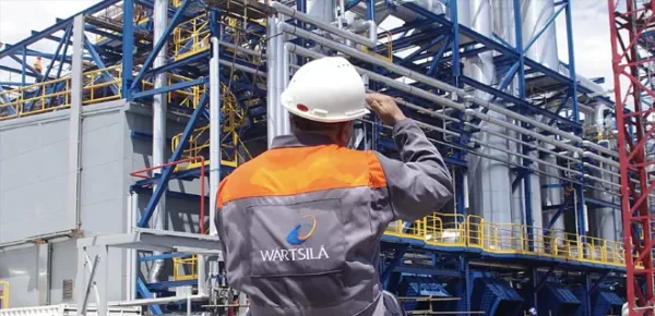A worker in a white hard hat and Wartsila uniform stands in front of an industrial facility with metal structures and pipes, facing away from the camera.