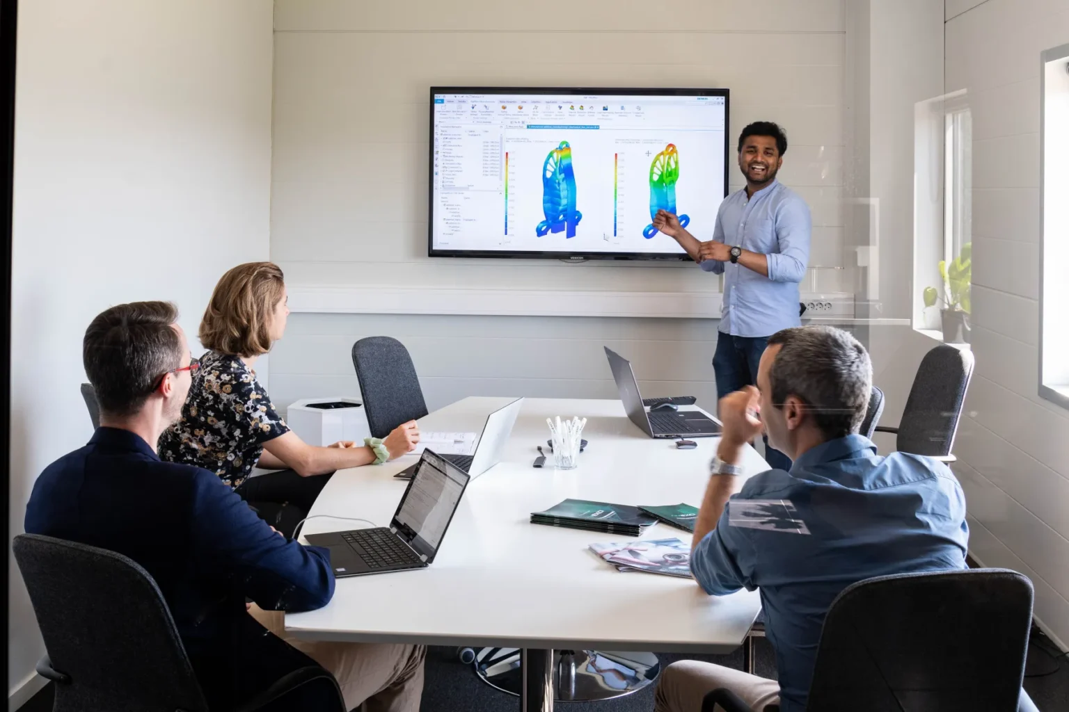 A man stands at the front of a conference room, pointing to a screen displaying colorful charts or simulations, while three colleagues sit at a table with laptops and notebooks, watching his presentation.