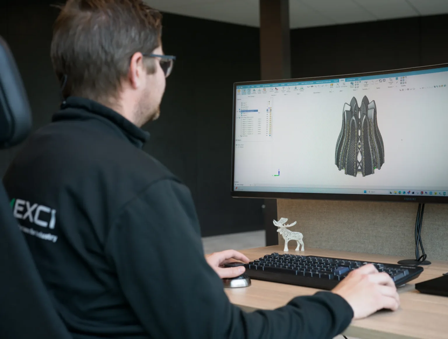 A person wearing glasses and a black jacket sits at a desk, using a computer with a large monitor displaying a 3D architectural design. A small reindeer figurine is placed in front of the monitor on the desk.
