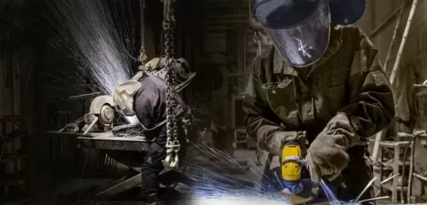 Two workers wearing protective gear use tools to grind and weld metal parts in an industrial workshop, with sparks flying around them. The background is dimly lit, highlighting the focused activities.