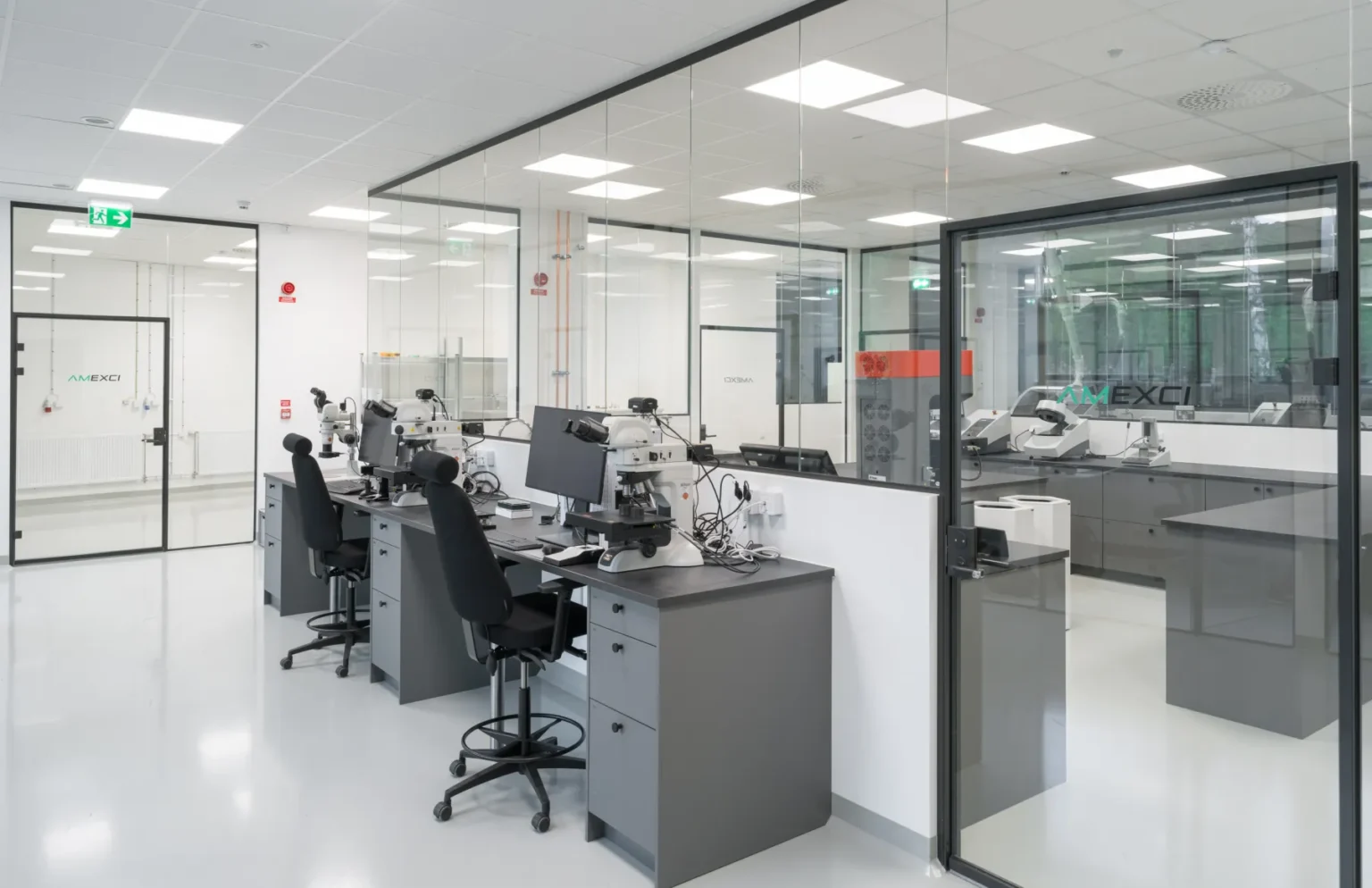 A clean, modern laboratory with glass walls, several desks equipped with microscopes, black office chairs, and computers. The space is well-lit with overhead lights and has a minimal, organized appearance.