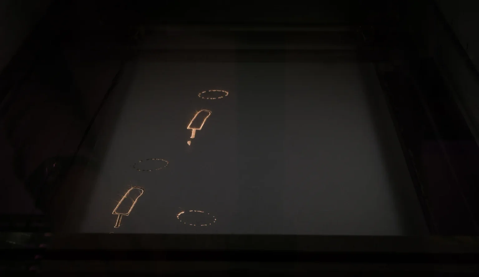A dark surface displays several illuminated outlines, including popsicle shapes and dotted circles, likely created by a laser or similar technology.