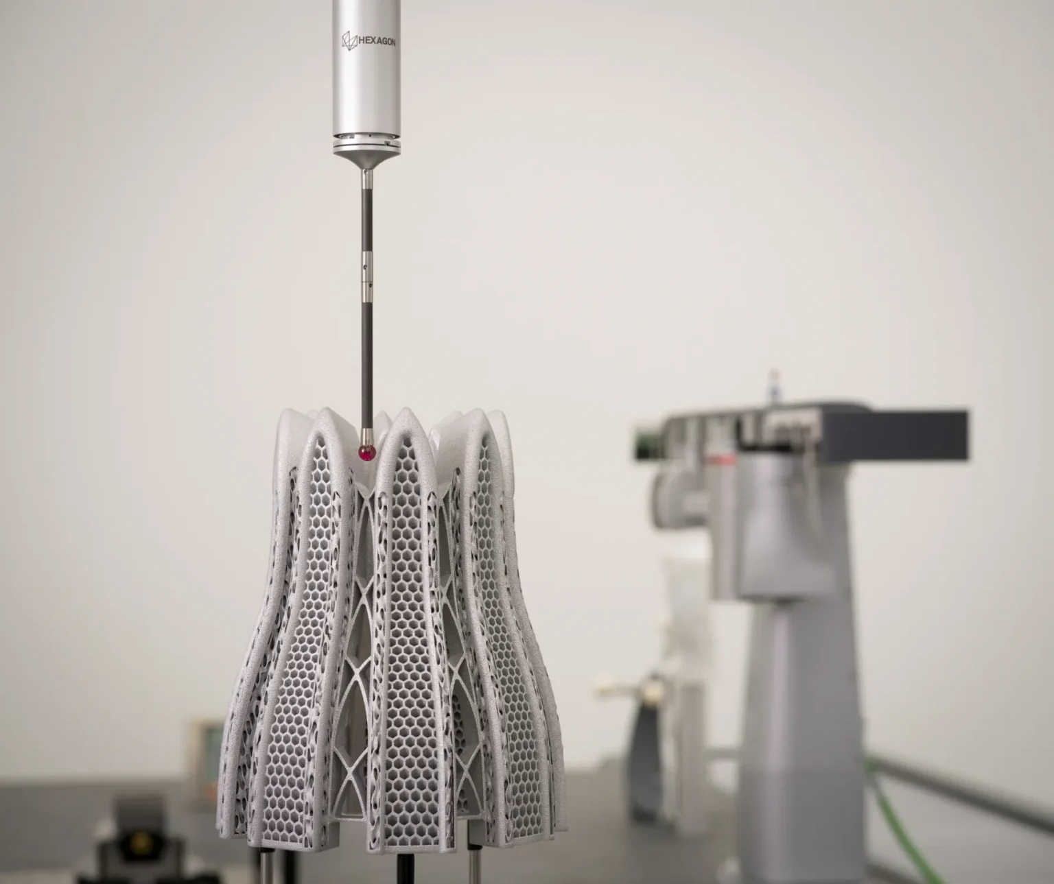 A coordinate measuring machine probe inspects a complex, lattice-structured metal part in a lab setting, with blurred measurement equipment visible in the background.