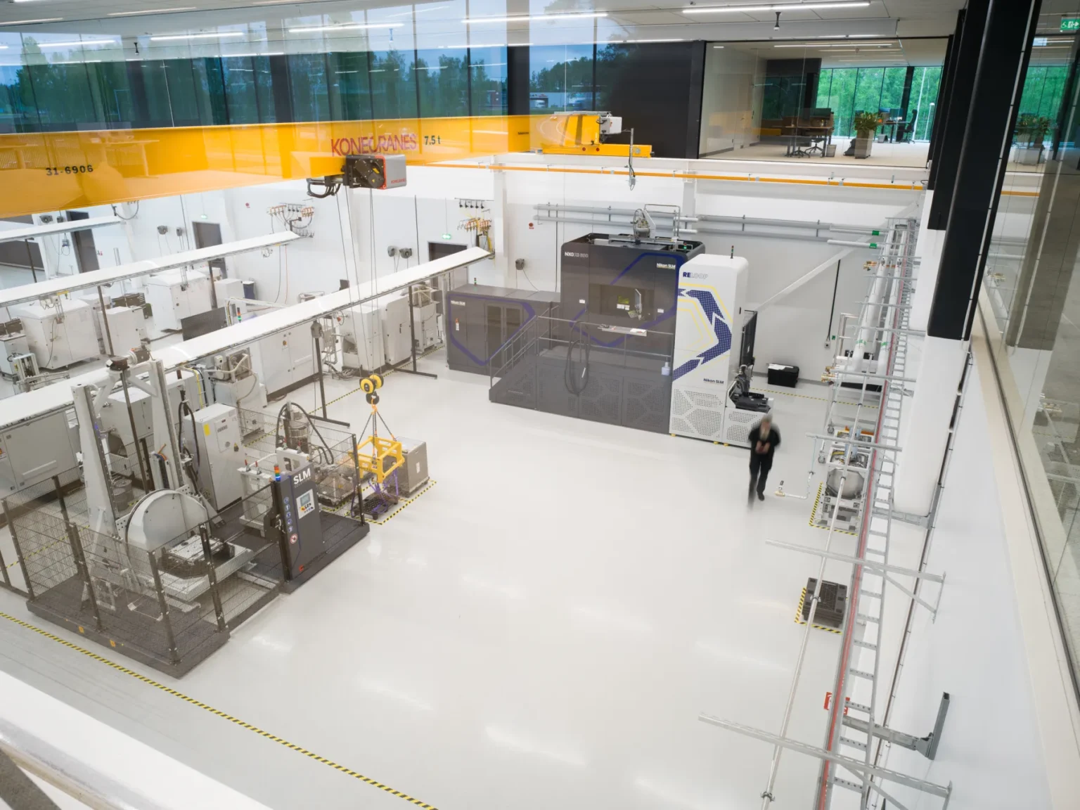 A spacious, modern industrial facility with various large machines, overhead cranes, workstations, and a person walking on the clean, white floor. The area is well-lit with natural and artificial light.