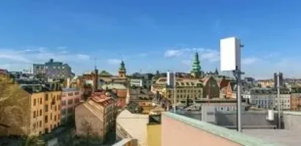 A cityscape view under a clear blue sky, with colorful buildings, church towers, and rooftops visible. In the foreground, there is a white rectangular antenna or device mounted on a rooftop.