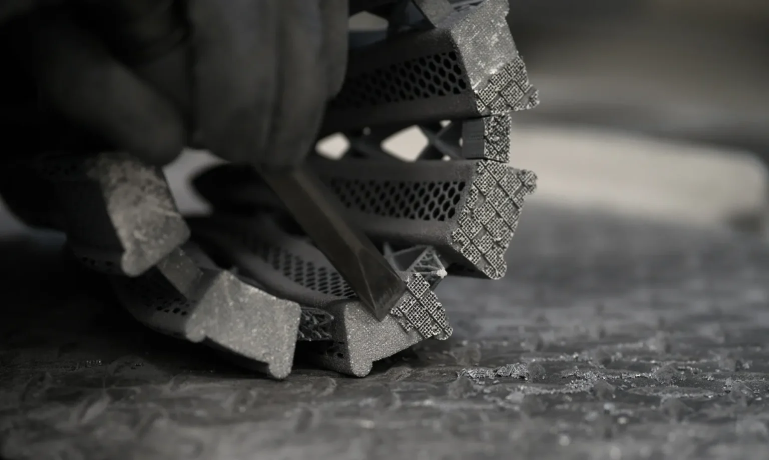A gloved hand uses a tool to carve or clean a detailed, lattice-patterned, metallic object resting on a textured surface.