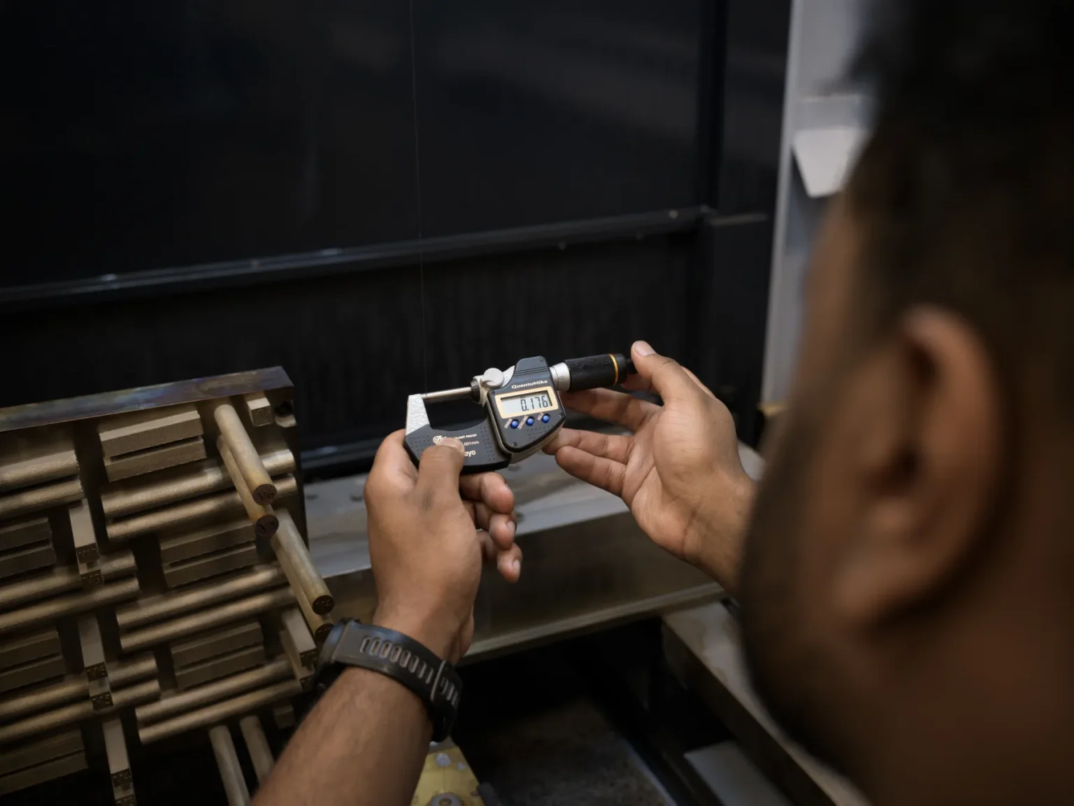 A person uses a digital micrometer to measure an object in a workshop or industrial setting, with wooden rods visible on the left side of the image.