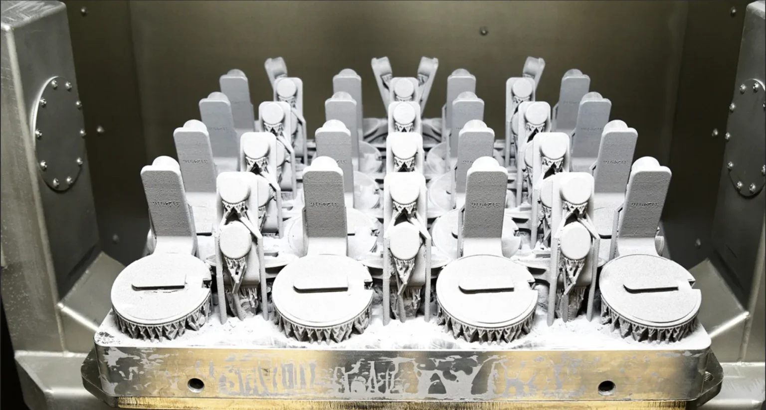 A tray filled with uniformly arranged, metallic 3D-printed machine parts covered in white powder, set inside an industrial printer or manufacturing chamber.