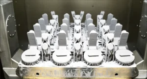 A tray filled with uniformly arranged, metallic 3D-printed machine parts covered in white powder, set inside an industrial printer or manufacturing chamber.