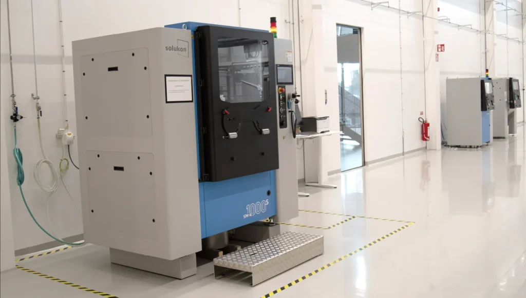 A large industrial machine with a blue and gray exterior stands in a clean, white room with other similar machines in the background. A metal step is placed in front, and safety markings are visible on the shiny floor.