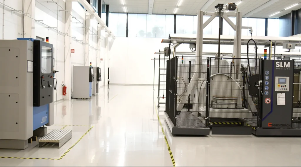 A clean, modern industrial facility with large 3D printing machines and equipment arranged on a shiny white floor, marked by yellow-black safety tape. The space is well-lit with large windows along the back wall.