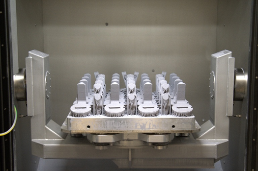 A metal 3D printer build plate holds multiple identical metal parts arranged in rows, inside the printers chamber. The parts appear unfinished with support structures attached.